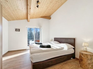 Une chambre avec un plafond en bois, un lit avec une literie blanche, une table de chevet avec une lampe et une fenêtre donnant sur des arbres.