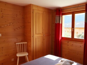 A room with wooden walls, a bed, a chair, and a window with curtains. - Petit Nid Douillet au Coeur de Salvan by Interhome (Salvan)