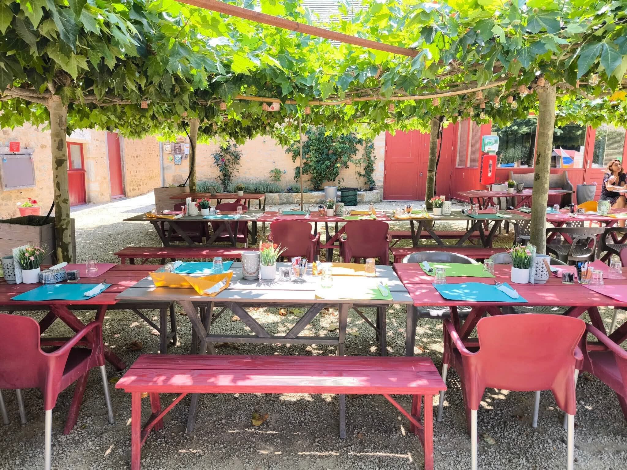 An outdoor dining area with tables and chairs, a pergola overhead, and a brick wall in the background.