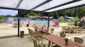 Un patio al aire libre con mesas y sillas, un espacio de estar cubierto y un parque infantil al fondo.