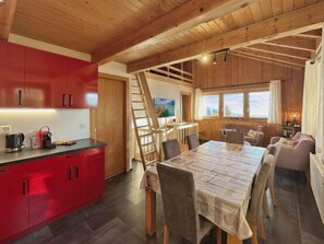 Eine moderne Küche mit roten Schränken, ein Essbereich mit großem Tisch und Blick ins Freie durch die Fenster.