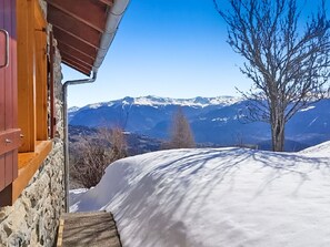 Eine Holzhütte mit einer Steinmauer, einem verschneiten Hang und einem Bergpanorama im Hintergrund.