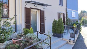 A building with a front door, brown shutters, and a small outdoor seating area with chairs and potted plants. - Affittacamere Brenna (San Fermo della Battaglia)