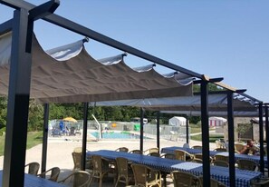 An outdoor patio with tables and chairs, a pool in the background, and a covered area with a view of the pool.