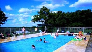 Piscina com pessoas na água, cercada por uma vedação e árvores ao fundo.