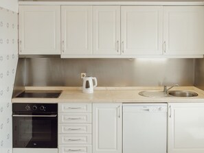 Una cocina con alacenas blancas, un lavamanos de acero inoxidable, una tetera, una estufa y un lavavajillas.