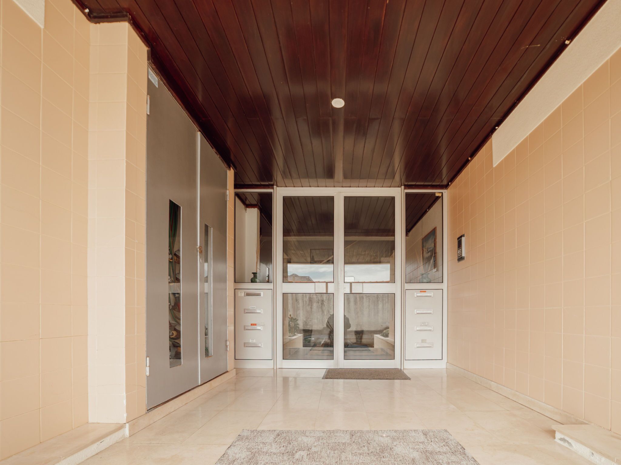 A hallway with a wooden ceiling, beige walls, and a glass door leading to a room with a desk and drawers.