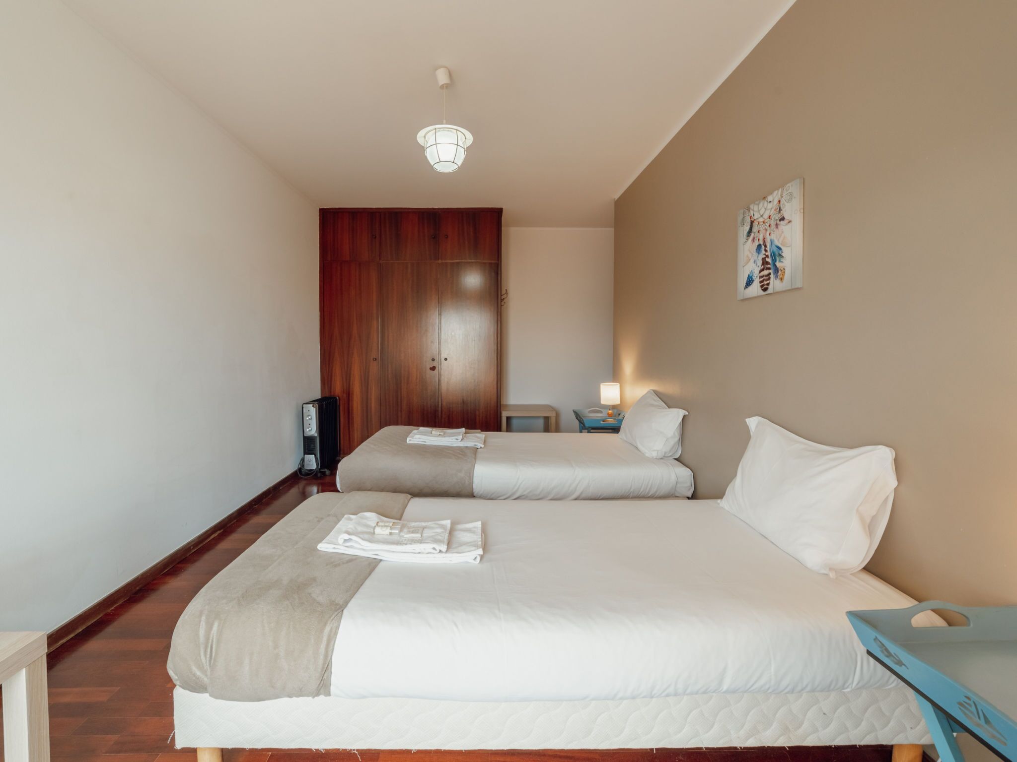 Habitación de hotel con dos camas, un armario de madera, una mesita de noche con lámpara y una obra de arte en la pared.