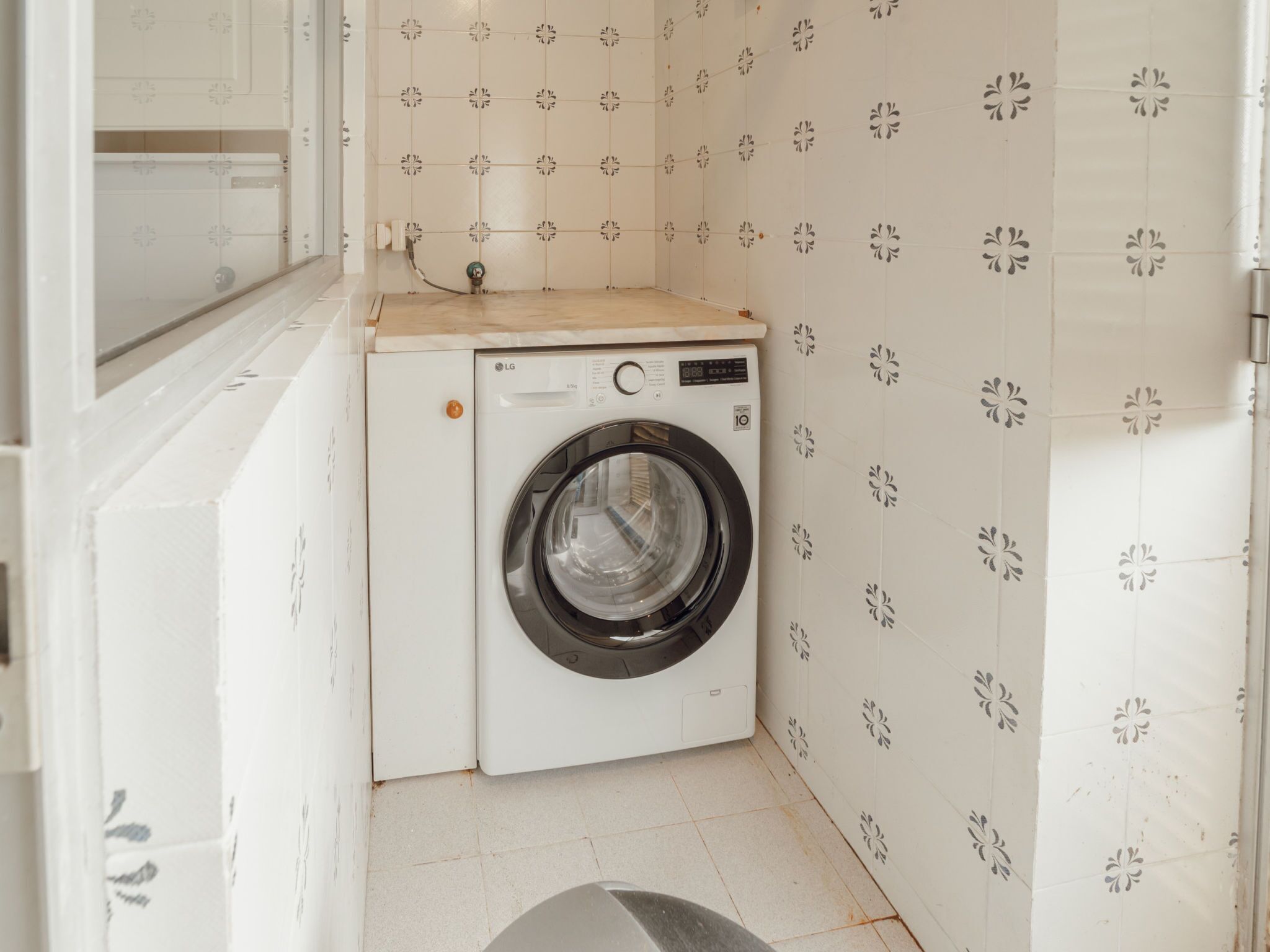 Máquina de lavar compacta e branca numa lavanderia pequena, com azulejos estampados nas paredes.