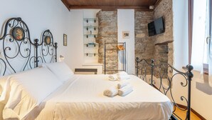 Una cama bien hecha con sábanas blancas y toallas enrolladas, una pared de piedra, un televisor montado y un techo de madera.