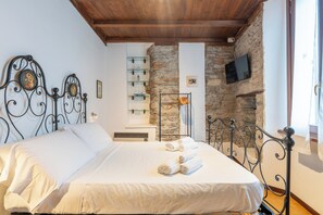 A neatly made bed with white linens and rolled towels, a stone wall, a mounted TV, and a wooden ceiling. - Historic Borgo Lake Como | Campidoglio 5 (Lenno)