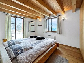 Ein Schlafzimmer mit Holzbalkendecke, einem Bett mit gemusterten Betttextilien, zwei Fenstern mit Vorhängen und Blick ins Freie.