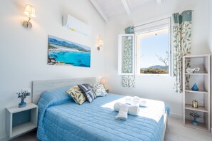A bedroom with a bed, pillows, a nightstand, a wall-mounted lamp, a painting, a window with curtains, and a shelf with decorative items. - Affittimoderni Castelsardo - Panorama (Castelsardo)