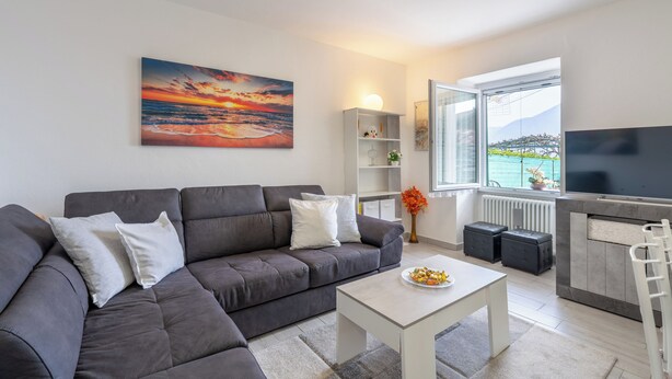 A modern living room with a grey sofa, a flat-screen TV, a painting, and a view of the sea. - The Amber - Tremezzo Lake Como (Tremezzo)