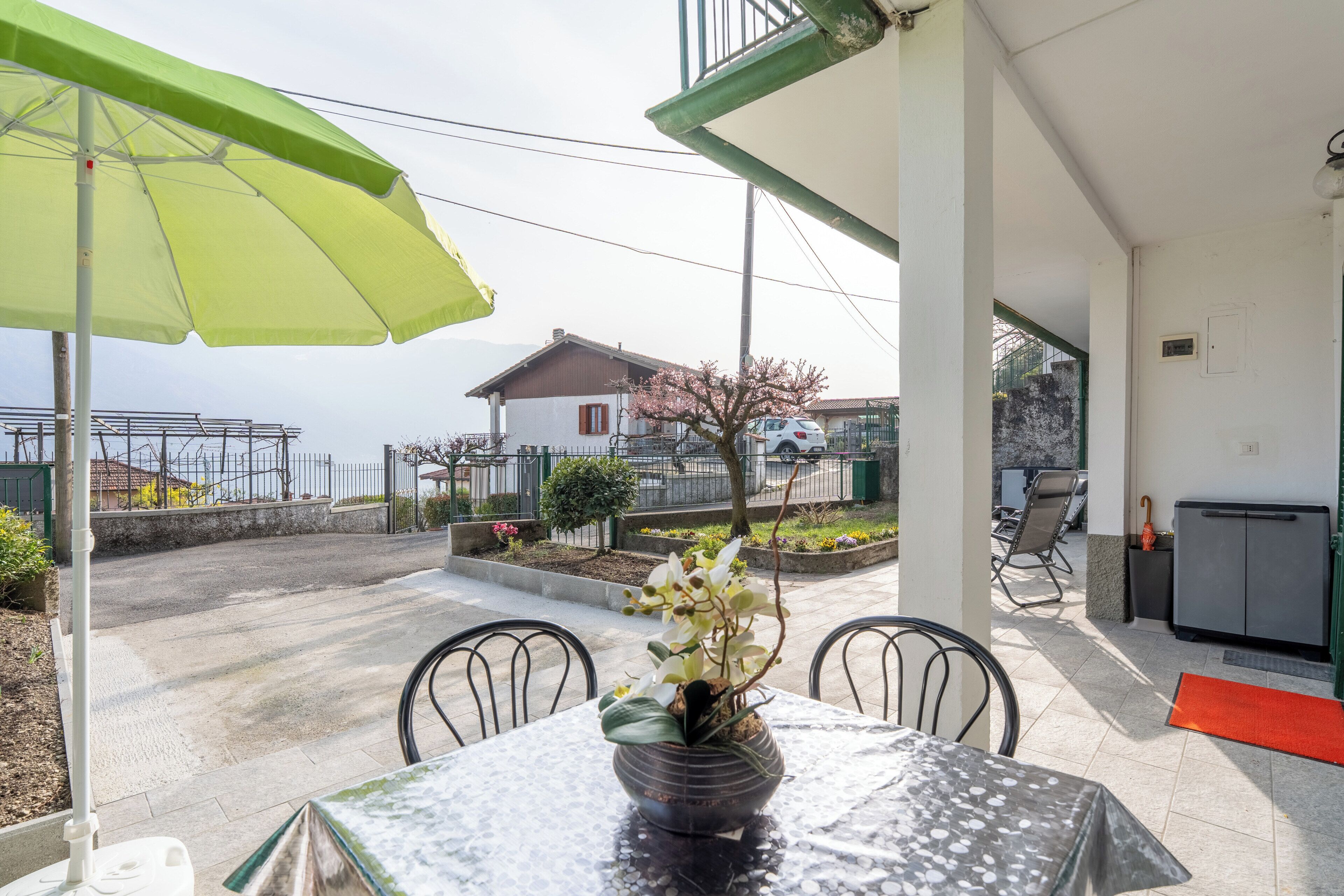 A patio with a table, chairs, a green umbrella, and a view of a house and garden. - The Amber - Tremezzo Lake Como (Tremezzo)