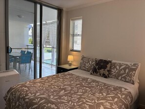A bedroom with a bed, a nightstand, a lamp, a window with blinds, and a view of a balcony with chairs. - Merivale Stay in South Brisbane two Beds two Baths one Parking (Brisbane)