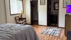 A bedroom with a bed, a desk, a chair, a window with blinds, a door leading to a bathroom, and a television on a stand. - Hayward Riverside Rental (Hayward)