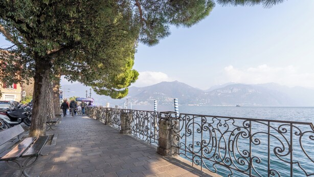 Eine gepflasterte Uferpromenade mit Bänken, einer Brüstung und Blick auf das Meer und die Berge.