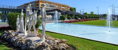 Una fontana con sculture in un'area verde curata, un ponte e un edificio sullo sfondo.