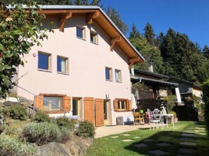 Ein zweistöckiges Haus mit geneigtem Dach, Holzläden und einer kleinen Terrasse mit Gartenmöbeln.