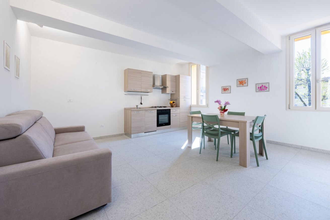 Un salon moderne avec un canapé, un coin repas comprenant une table et des chaises, une cuisine équipée de meubles et d’appareils électroménagers, et une grande fenêtre laissant entrer la lumière naturelle.