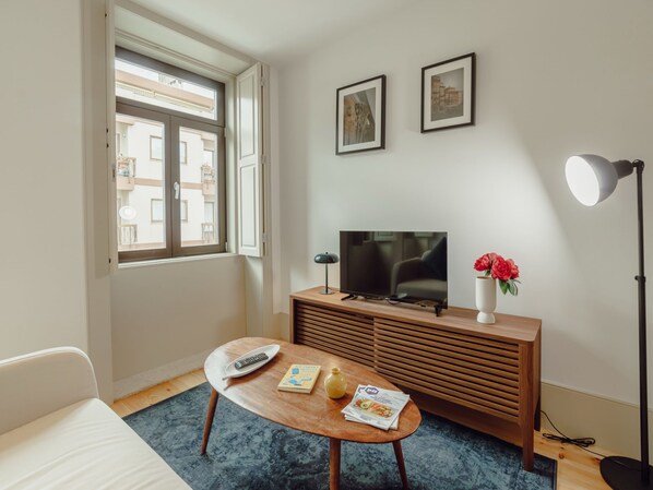 A room with a television on a wooden cabinet, a remote control, a book, and a vase with flowers on a small table. - Apartment  by Interhome (Porto)
