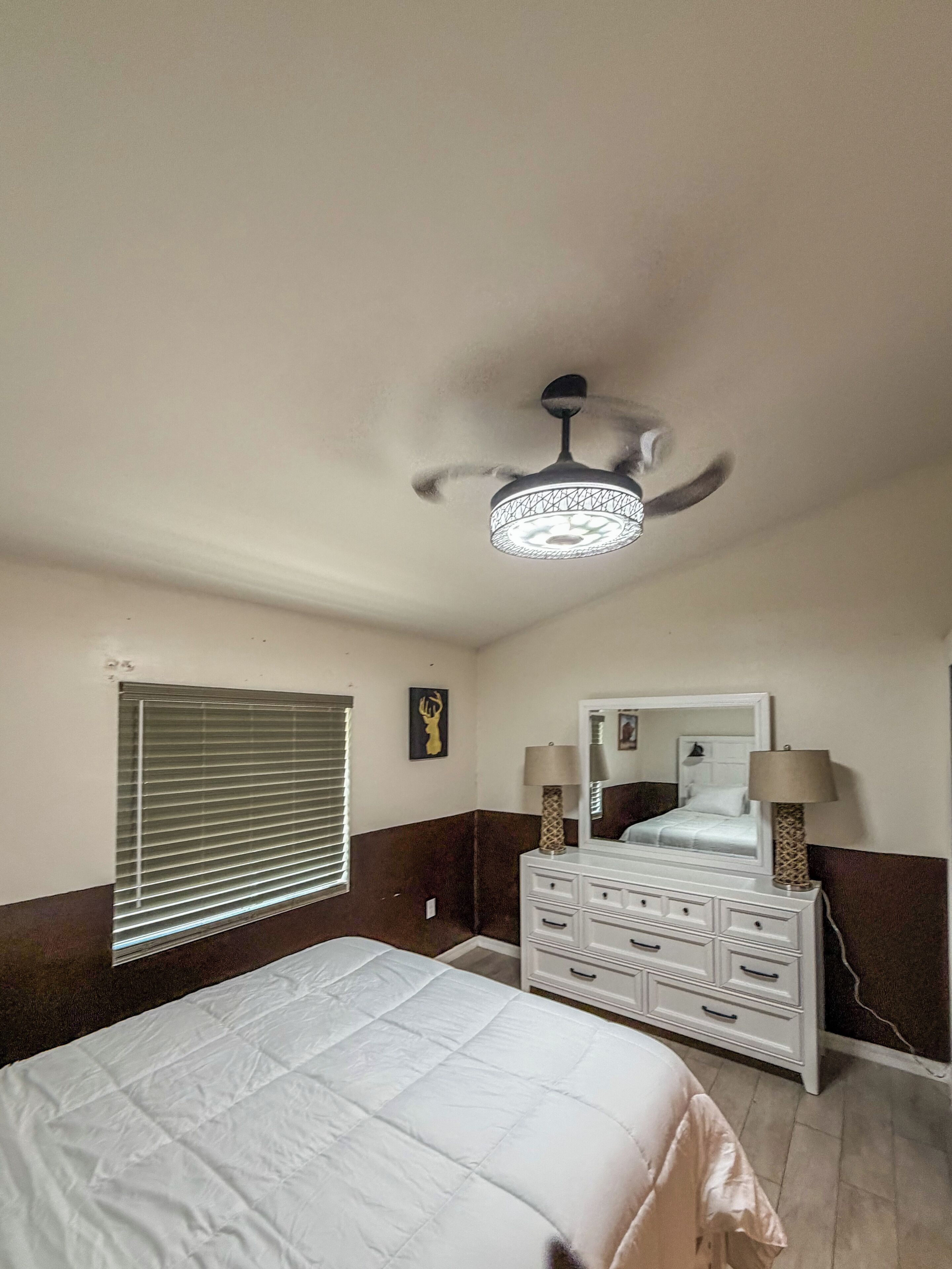 A bedroom with a bed, dresser, mirror, ceiling fan, and window with blinds.