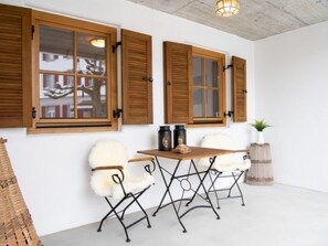 A room with a white wall, wooden shutters on windows, a white chair, a table with a basket, and a potted plant.