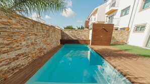 Ein rechteckiger Pool mit umlaufender Holzterrasse, vor einer Ziegelwand und einem Gebäude mit Balkonen.
