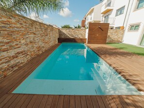 Ein rechteckiger Pool mit umlaufender Holzterrasse, vor einer Ziegelwand und einem Gebäude mit Balkonen.