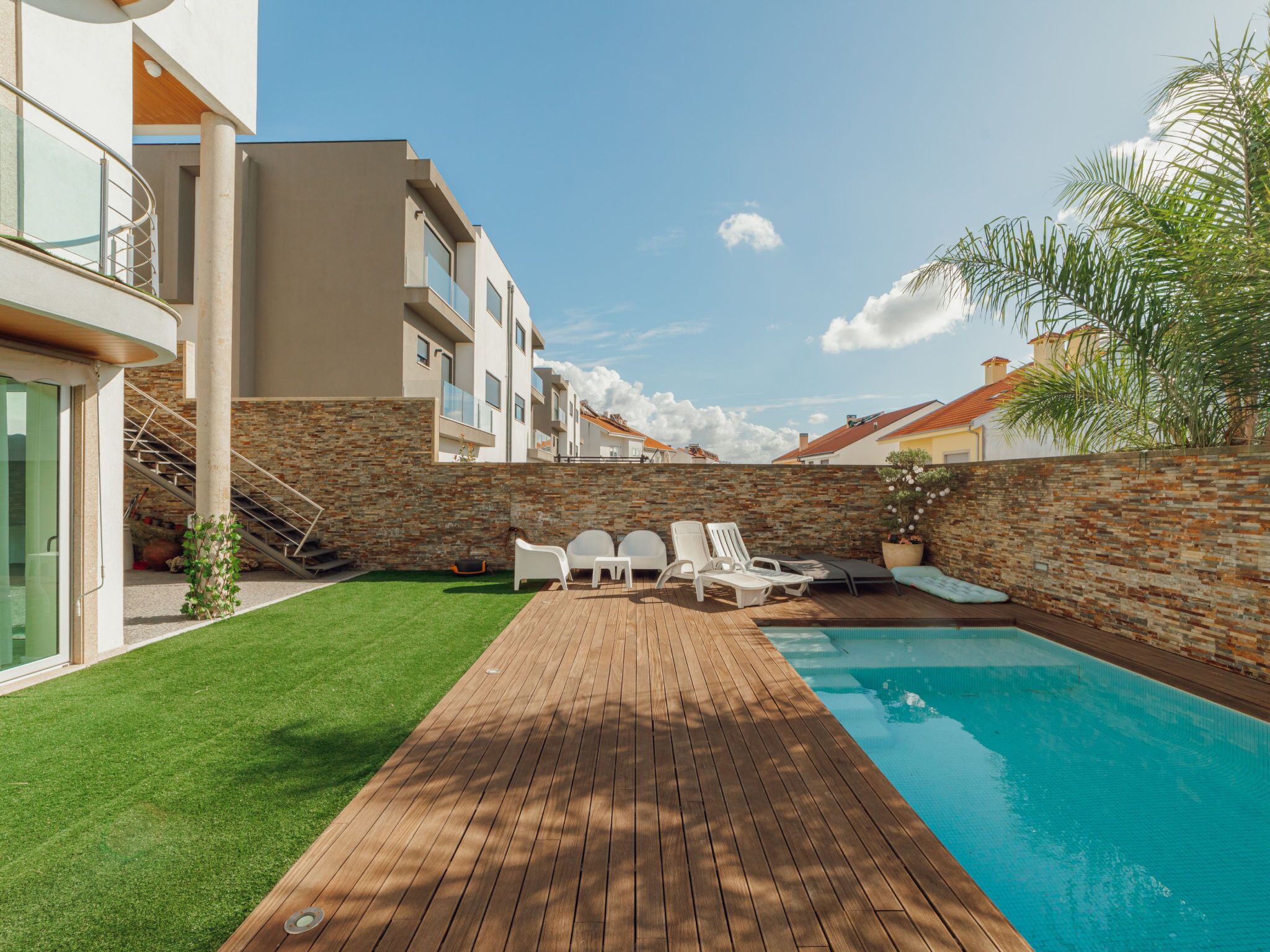 A modern outdoor pool area with a wooden deck, lounge chairs, and a stone wall.