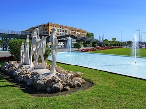 Ein Brunnen mit Skulpturen in einer angelegten Grünfläche, eine Brücke und ein Gebäude im Hintergrund.