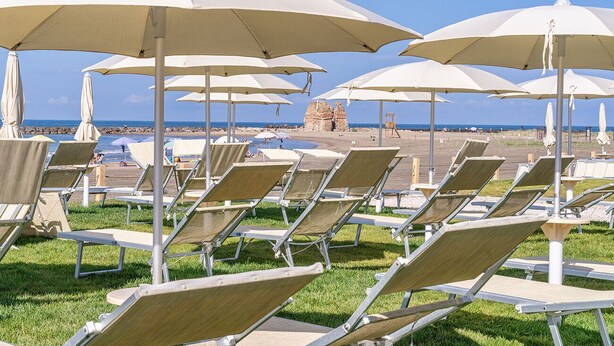 Rows of sun loungers with attached umbrellas, a clear sky, and a rocky outcrop in the background. - Gotha Beach Srl - Casetta Lavanda (Ladispoli)