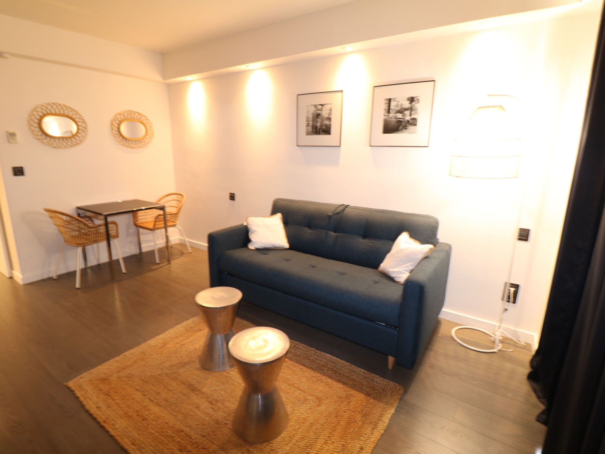 Ein modernes Wohnzimmer mit einem dunkelblauen Sofa, zwei weißen Kissen, einem kleinen Holztisch und einem gewebten Teppich. Es gibt zwei Korbstühle, einen Esstisch und zwei an der Wand befestigte Spiegel. Der Raum zeichnet sich durch ein aufgeräumtes, minimalistisches Design und einen hellen Boden aus.