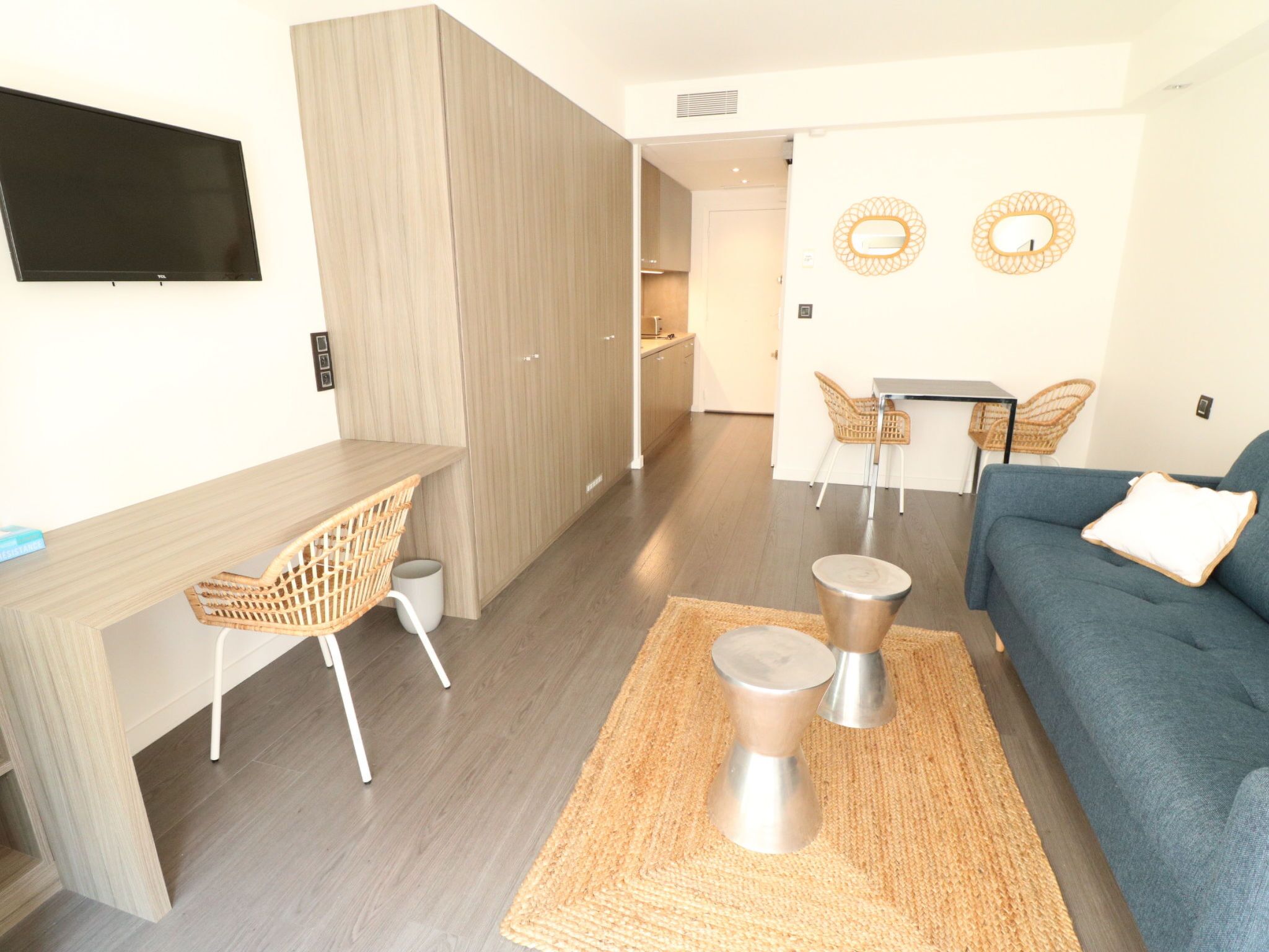 Un salon moderne avec un canapé, une table à manger et des chaises, un téléviseur fixé au mur, et un coin cuisine à l’arrière-plan.