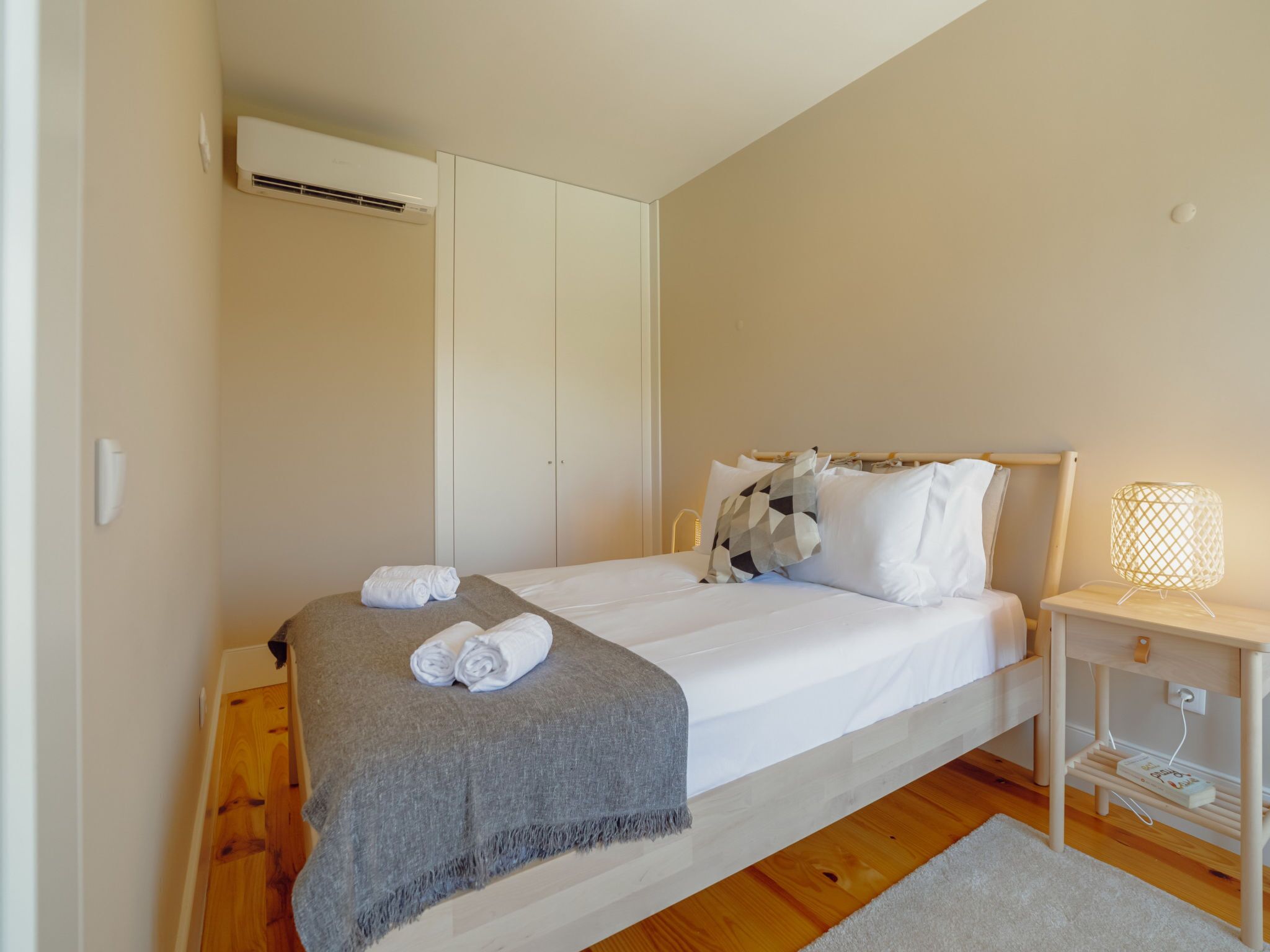 A bedroom with a bed, a bedside table with a lamp, a grey throw blanket, and rolled towels.