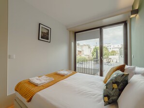 Ein Hotelzimmer mit einem Bett, einem Balkon mit Blick auf Gebäude und einem gerahmten Bild an der Wand.