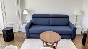 Una sala de estar con un sofá azul oscuro, dos mesitas de noche blancas con lámparas, una mesa redonda de madera y una alfombra blanca.