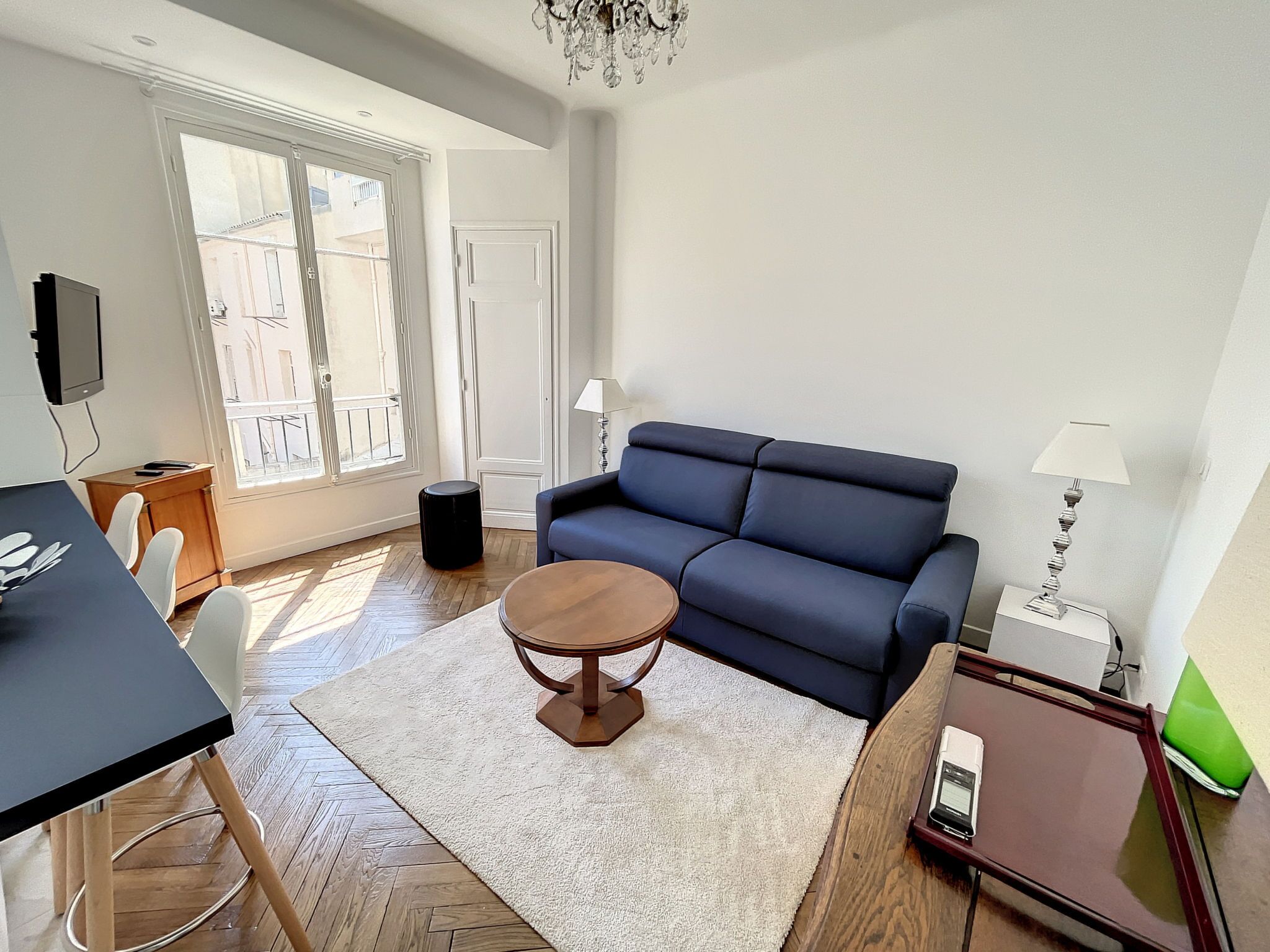 A living room with a sofa, a round table, a TV mounted on the wall, and a window with a view of buildings.