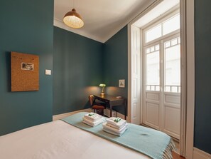 Une chambre d’hôtel avec un lit, un bureau, une chaise, une fenêtre et un panneau d’affichage en liège au mur.