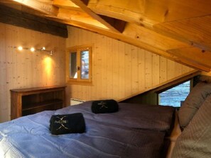 Ein Zimmer in einer Holzhütte mit schrägverlaufender Decke, einem Bett mit Kissen, einer kleinen Holzbank und einem Fenster mit Blick.