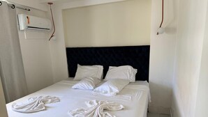 Una habitación de hotel con cama, ropa de cama blanca, cabecera oscura y unidad de aire acondicionado.