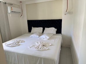 A hotel room with a bed, white linens, a dark headboard, and an air conditioning unit. - Beachfront apartment in Recreio (Rio de Janeiro)