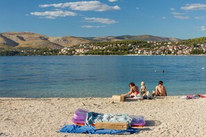 Een strand met mensen die zitten en zonnebaden, een heldere lucht en een meer met een stad op de achtergrond.