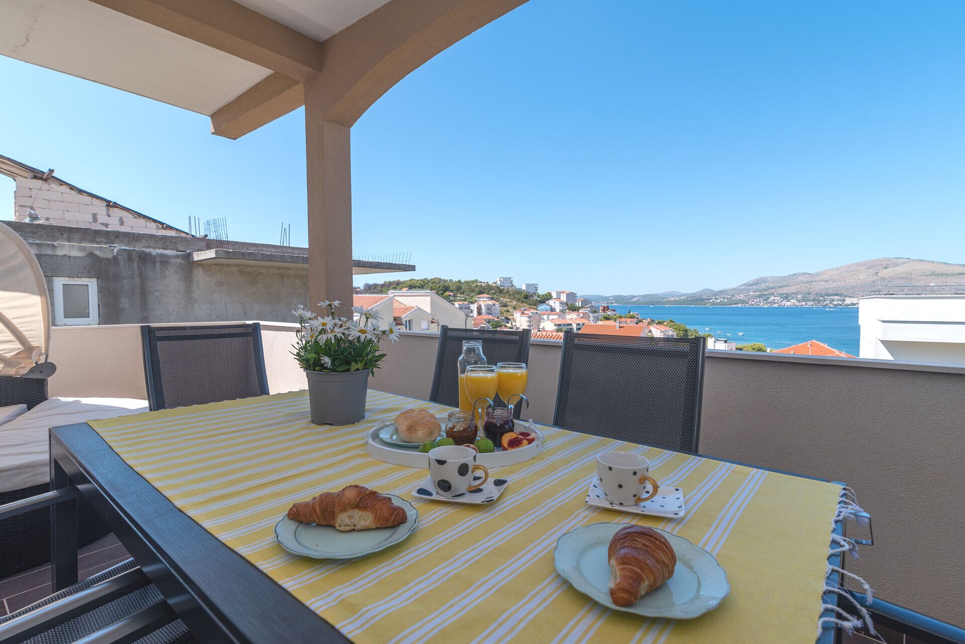 Un balcón con una mesa dispuesta para el desayuno, que incluye cruasanes, una bandeja de fruta y vistas al mar.
