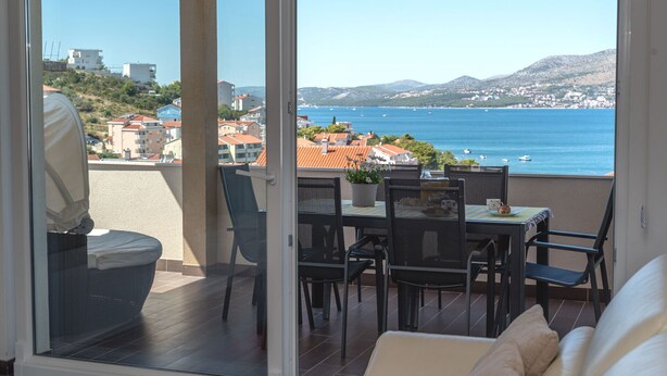 A balcony with a table and chairs, overlooking a coastal view with buildings and mountains.