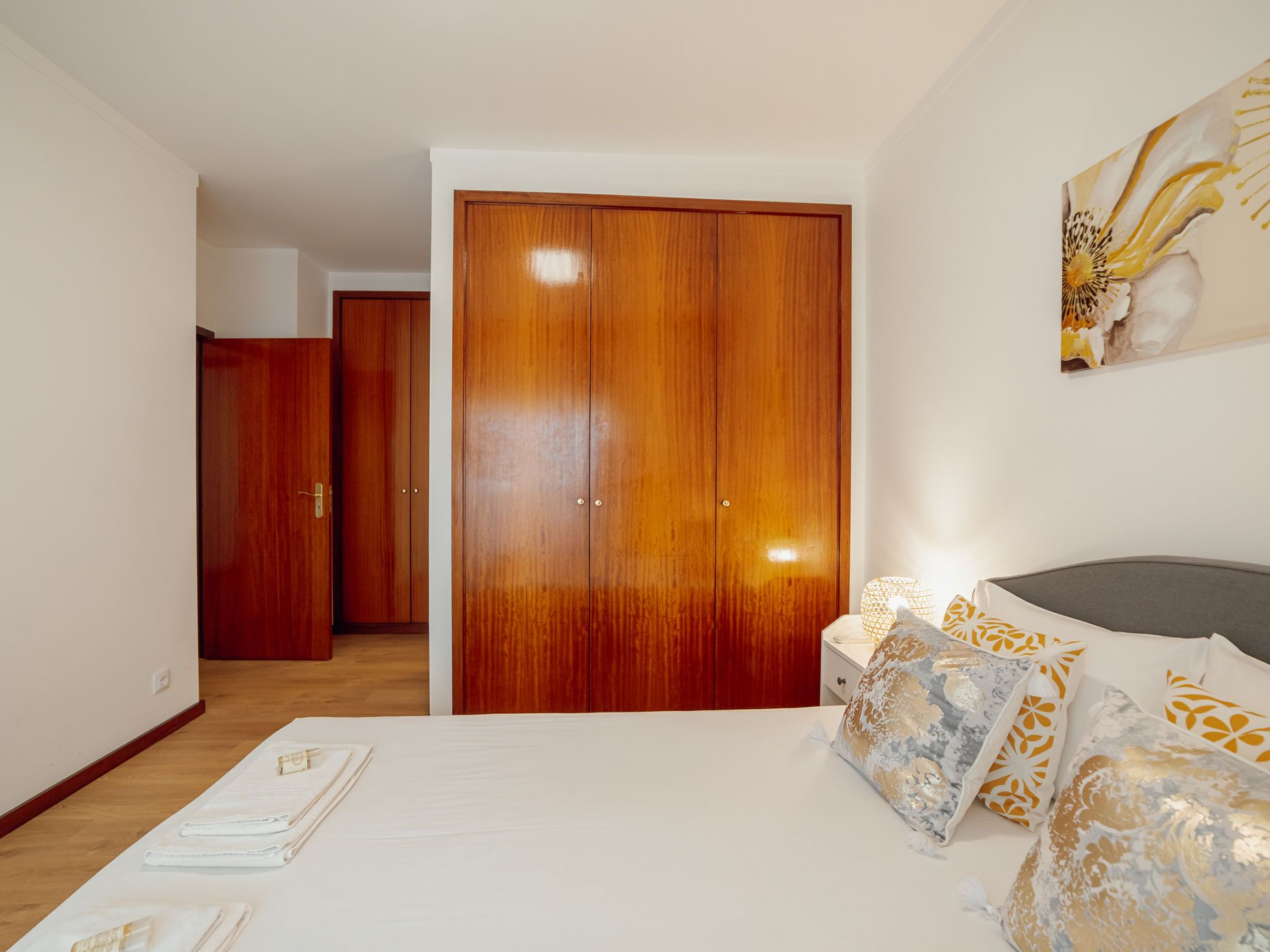 Un dormitorio con un armario grande de madera, una cama con almohadones estampados y una mesita de noche con una lámpara.