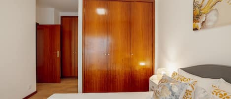 Un dormitorio con un armario grande de madera, una cama con almohadones estampados y una mesita de noche con una lámpara.