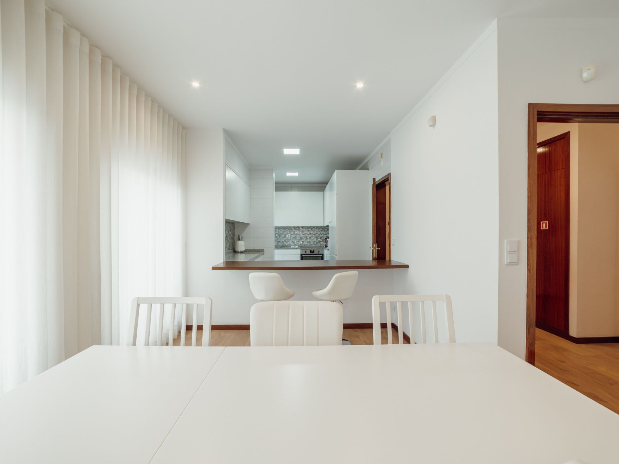 A modern dining area with a white table and chairs, a kitchen in the background, and a wooden door on the right.