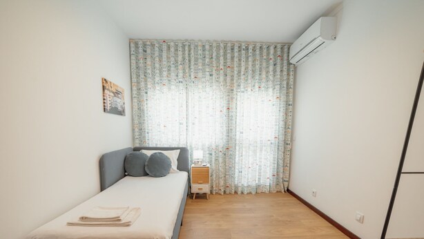 Un dormitori amb un llit, una tauleta de nit, una làmpada, una finestra amb cortines estampades i un aparell d'aire condicionat.